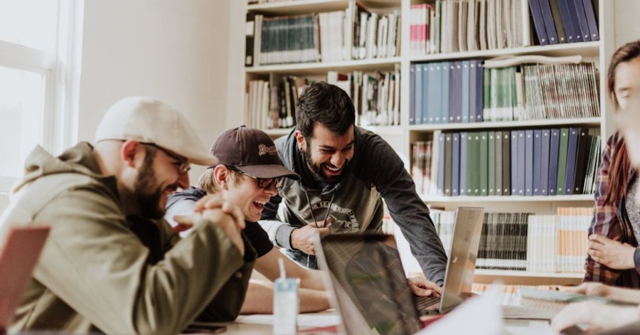 Men in a library, laughing while watching something on a laptop